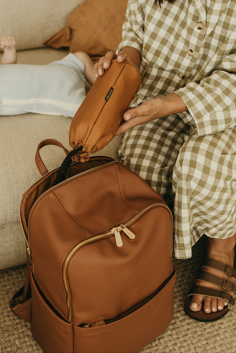 Woman sitting on a couch with a baby lying down, opening a brown backpack.