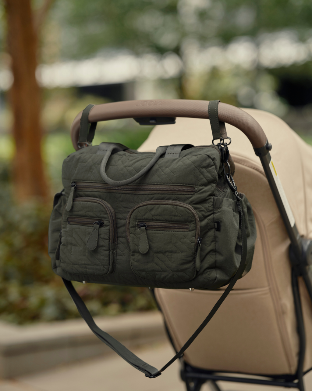 Green diaper bag attached to a beige stroller with a blurred outdoor background