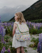 Child with a floral backpack standing in a field of lupin flowers with mountains in the background
